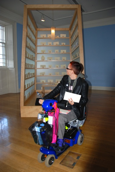 Colour photograph of me on my mini scooter in front of the letter writing booth in Dean Gallery, holding an envelope addressed to Lee Mingwei.