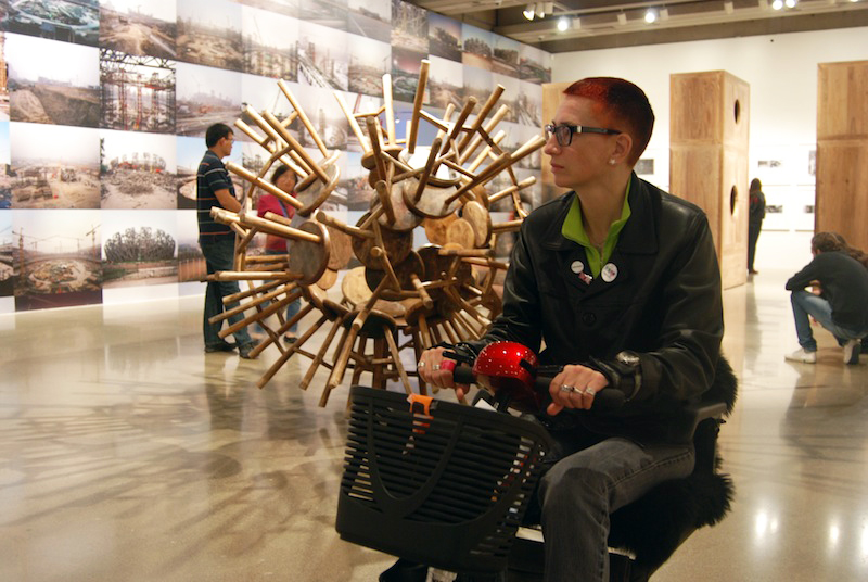 Ju sitting on a red mobility scooter in front of a sculpture made from wooden stools