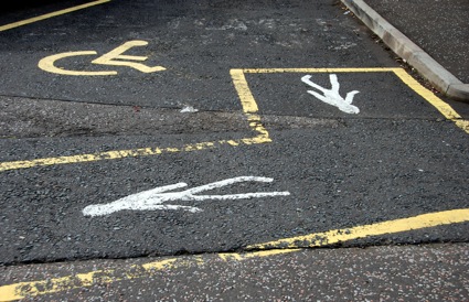 Colour photograph of a 'disabled' parking space, where the wheelchair user symbol in the space itself is replaced by walking figures in the marked path leading from the space.