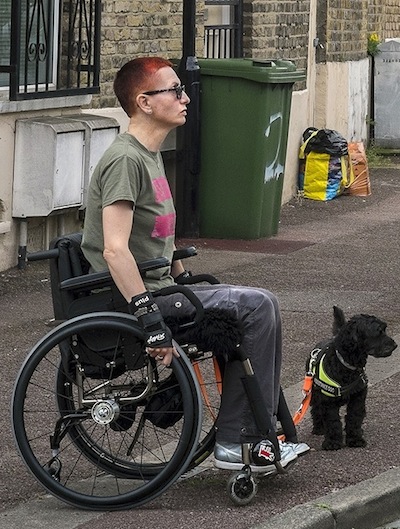 Colour photograph of a white woman with short red hair, sitting in the street in a manual wheelchair accompanied by a small black dog in a hi-vis jacket