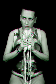 Black and white photo showing ju90, dressed in a her decorated spinal brace, a silver vest and black g-string, sitting holding her ballet shoes in front of her and looking into the distance with a determined expression on her face.