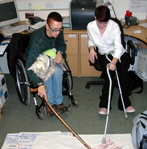 Colour photograph of two women sitting in an office, using their stick and crutches to point at entries on a list written on paper in front of them.