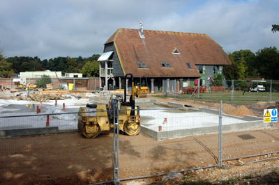 Colour photograph of a building site, showing foundations laid in concrete, with the Barn in the background