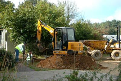 Colour photograph of a hole being dug next to a path, with two men working manually and one operating a small yellow mechanical digger and another machine in the background