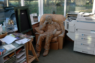 Colour photograph of a sculpture of a man slumped in an armchair, made of cardboard, set between a photocopier and a table laden with paperwork