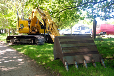 Colour photograph of a large yellow digger parked on a grass verge by the side of the lane.
