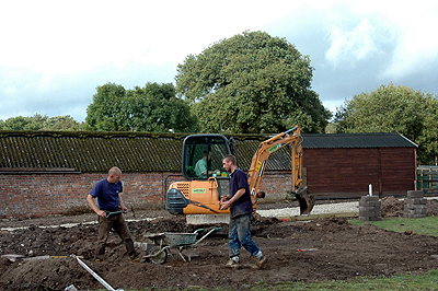 Colour photograph of three men digging the earth, two by hand and one in a small yellow mechanical digger, with farm buildings behind them