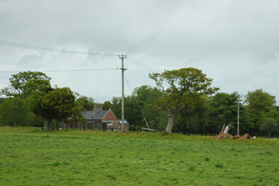 Colour photograph showing a grassy field in the foreground, with a brick and slate farm house buried in trees in the background. The sky is cloudy and grey.