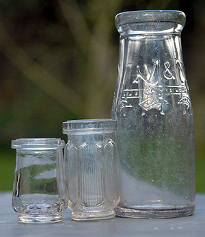 Close-up colour photograph of an old-fashioned glass milk bottle and two small glass jars, taken outdoors
