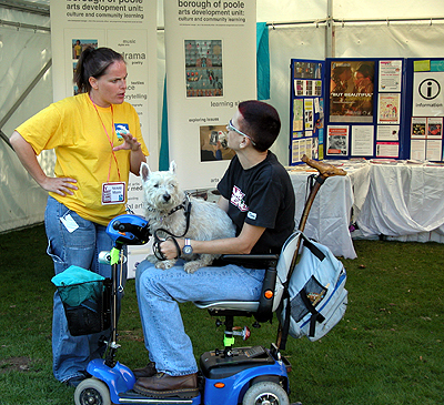 Colour photograph of Ju on her small blue mobility scooter with her dog on her lap, talking to a woman in a yellow t-shirt in front of a marquee displaying information material