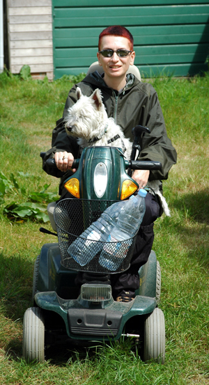 Photograph of the artist, dressed in rain mac and sunglasses, sitting on a large green scooter with a Westie on her lap