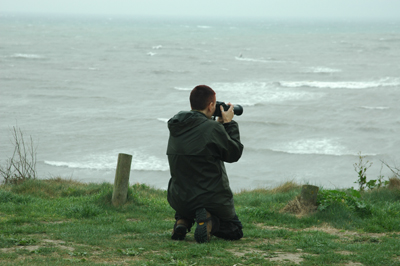 Colour photograph of the artist kneeling down on a cliff top and holding a camera, with grey waves in front of her
