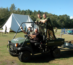 Colour photograph of four men dressed in army fatigues, riding a small truck, with a marquee being taken down in the field behind them