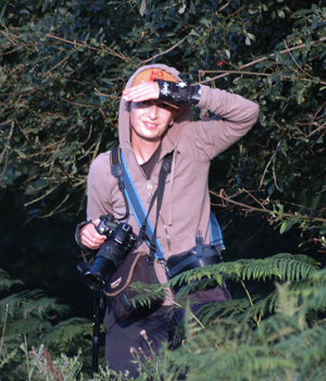 Colour photograph of me smiling into the sunshine from the undergrowth, hung with camera bags.