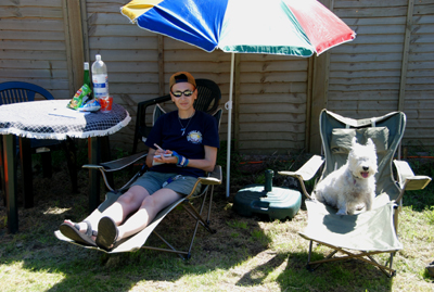 Colour photograph of Genie and I sitting on neighbouring green collapsible sun beds, with a parasol over us; I have a notebook and pen in my hands.