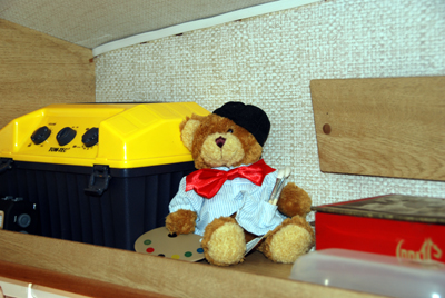 Colour photograph of a teddy bear dressed in an artist's smock and beret and carrying paints, sitting on the caravan shelf.