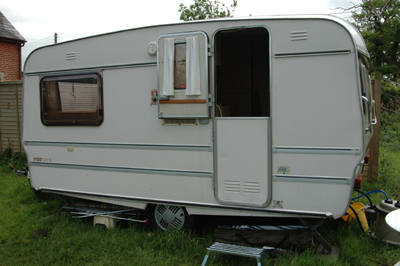 Close-up colour photograph of a white caravan on grass. It has a 'stable' door, and the top half is open while the bottom half is closed. Behind the caravan is a fence, and over it can be seen the end of the brick cottage wall.