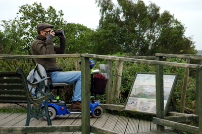 Colour photograph of the artist sitting on a small blue scooter on a wooden platform, taking photographs
