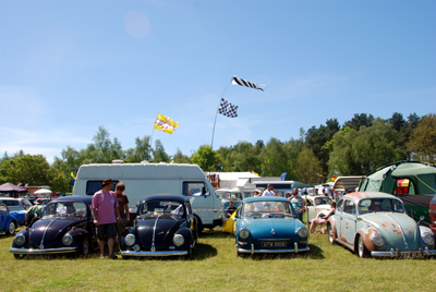 Colour photograph of VW Beetles and camper vans, crowds of people and flags flying against a blue sky.