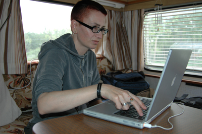 Colour photograph of Ju, wearing black-framed glasses and a grey zipped hooded top, sitting working at  her laptop at the caravan table. The colours of the caravan furnishings are orange and brown.