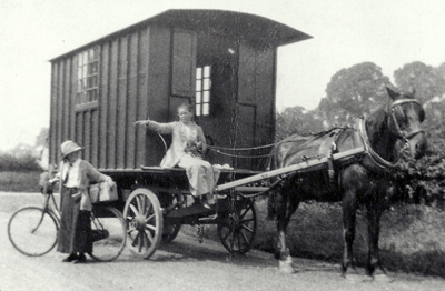 Black and white photograph of a woman in a long dress and a bun driving a horse-drawn caravan, talking to a woman on a bicycle
