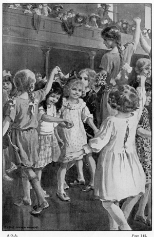 Black and white photograph showing young children folk dancing in a hall, with people looking on from a balcony.