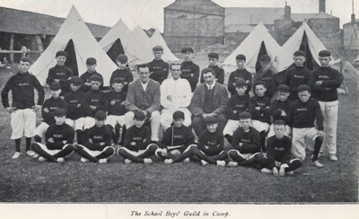 Black and white photograph of boys in uniform sitting in front of bell tents