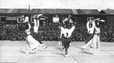 Black and white photograph of a group of men and women folk dancing in front of soldiers seated on the ground.