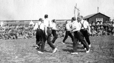 Black and white photograph of men sword dancing in front of soldiers seated on the ground.