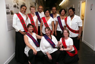 Colour photograph of a mixed group of ten adults, dressed in white tops and black trousers or skirts, and all wearing brightly coloured sashes over their shoulders embroidered with the names of folk dances.