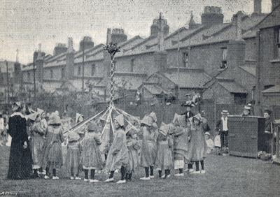 Black and white photo of children dressed in smocks and caps plaiting a maypole with the backs of terraced houses behind them.