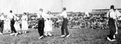 Black and white photograph of a group of men and women folk dancing in front of soldiers seated on the ground.
