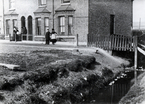 Black and white photograph of people in their homes, next to a ditch full of water
