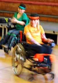 Colour photograph of two dancers, one dressed in green and clapping and one dressed in orange who is pivoting round.