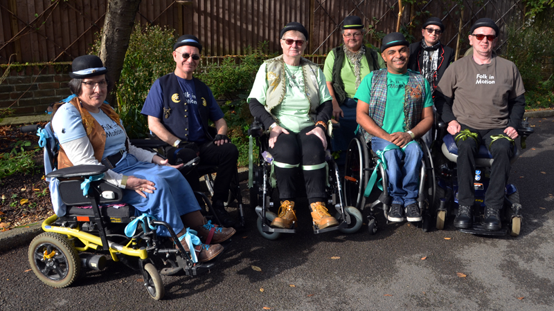 7 wheelchair users are sitting in a group outside dressed in coloured 'Folk in Motion' T-shirts, bowler hats and waistcoats.