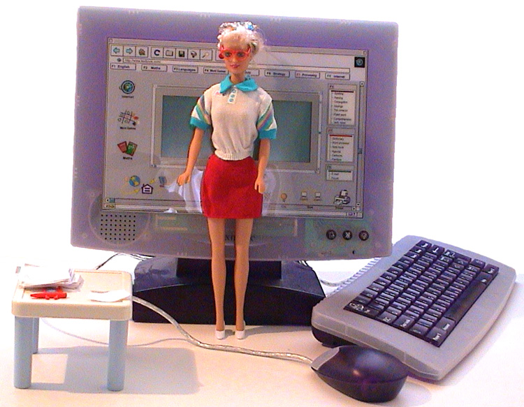 A fashion doll dressed in a skirt and blouse and red glasses faces front against a white background. Behind her are a child's computer toy and a doll's table on which rests pieces of paper and a plastic doll's pen.