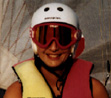 Head and shoulders shot of a smiling person in a white crash helmet, large pink visor and yellow and pink sleeveless jacket, in front of a white sail