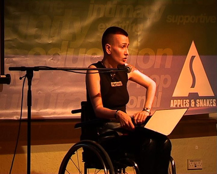 Photograph of ju90 performing in wheelchair, with Apples and Snakes banner behind