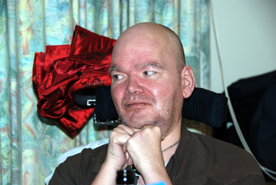 Colour photograph of a middle-aged man's shaved head against a wheelchair head rest on which a bright red satin dressing gown is resting. His hands are propped together under his chin, and behind him are turquoise patterned curtains.