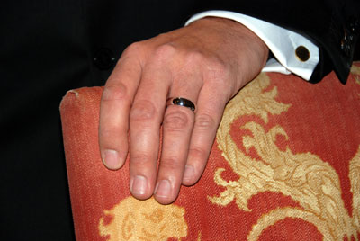 Colour photograph of a man's hand wearing a wedding ring, resting on a red and gold settee.