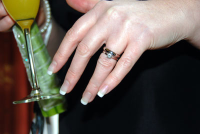 Colour photograph of a woman's hand wearing a wedding ring and engagement ring, with a champagne flute in the background.