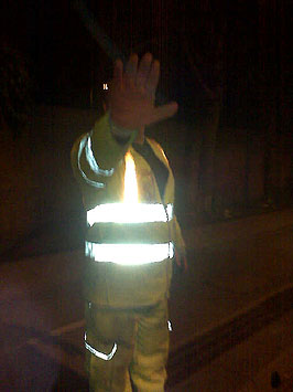 Colour photograph of a man holding his hand out over his face at night, and wearing reflective clothing.