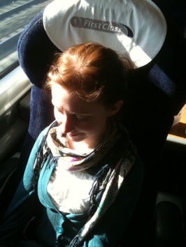 Colour photograph of a smiling young woman sitting in a first class train seat, with her naturally red hair gleaming in the sunlight.