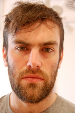 Colour photograph of the head of a young man, with dark hair and beard, wearing a grey t-shirt.