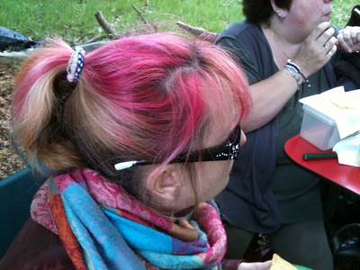 Colour photograph of the side view of a woman whose blond hair has been streaked pink and tied up behind her with a gingham hair band. She is wearing elaborate black-framed glasses and a multicoloured scarf.