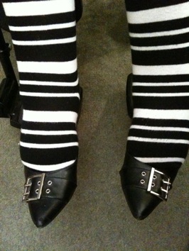Colour photograph of a seated woman's legs and feet in striped black and white tights and pointed black buckled shoes.