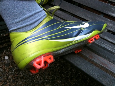 Colour photograph of a teenage boy's large foot in a lime green and orange football boot and grey sock against a garden bench.