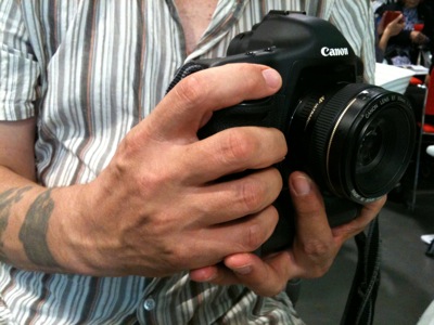 Colour photograph of a man's hands gripping a Canon camera against a white striped shirt. One wrist is tattooed.