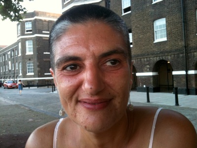 Colour photograph of the head and shoulders of a tanned woman with strong features and greying short hair, smiling against a background of riverside apartments.