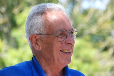 Colour photograph of the head and shoulders of a smiling older man with white hair and glasses, wearing a royal blue polo shirt.